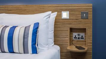 A bed with a striped pillow, a power outlet, and a phone charging on a wooden headboard.