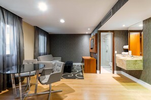 A modern hotel room with a glass dining table, two chairs, a sofa, a small wooden side table, a bathroom with a vanity, and a shower area. - Wyndham Sao Paulo Ibirapuera Convention Plaza Hotel (São Paulo)