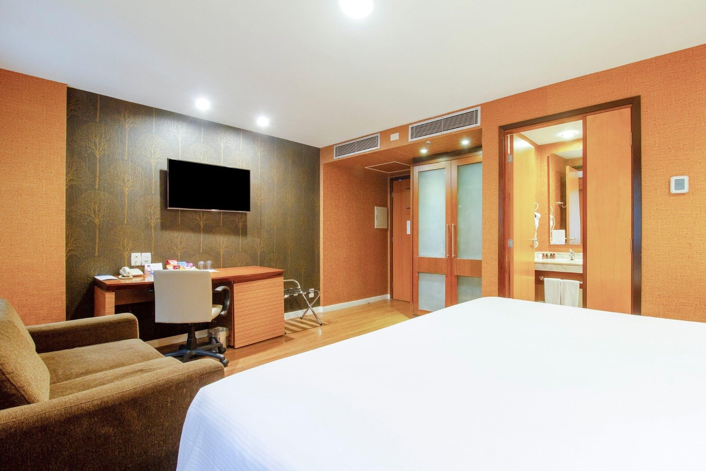 Habitación de hotel con cama, escritorio, silla, televisor y baño visible a través de una puerta abierta.
