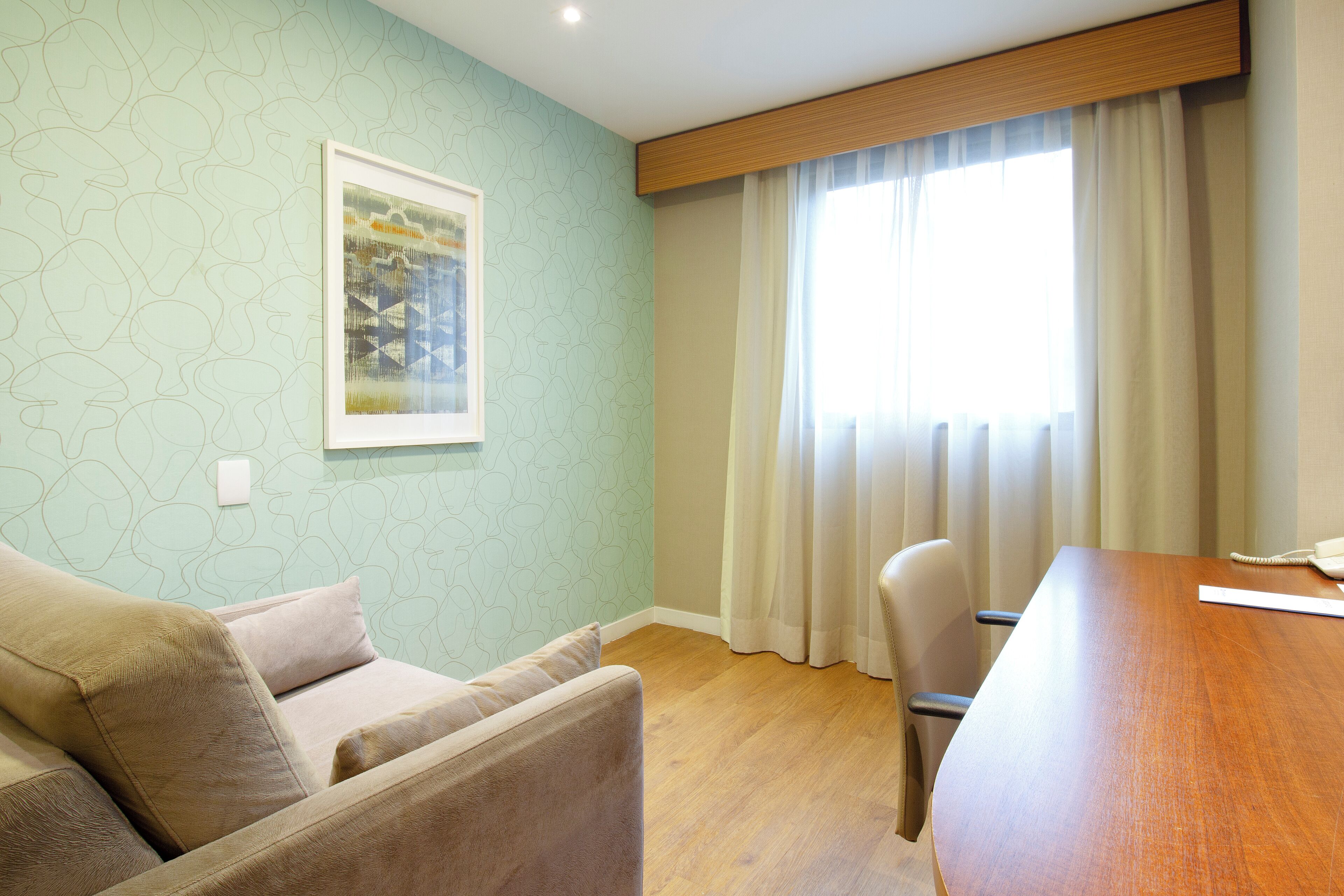 Una stanza con un tavolo di legno, un divano beige, un quadro appeso al muro e una finestra con tende.