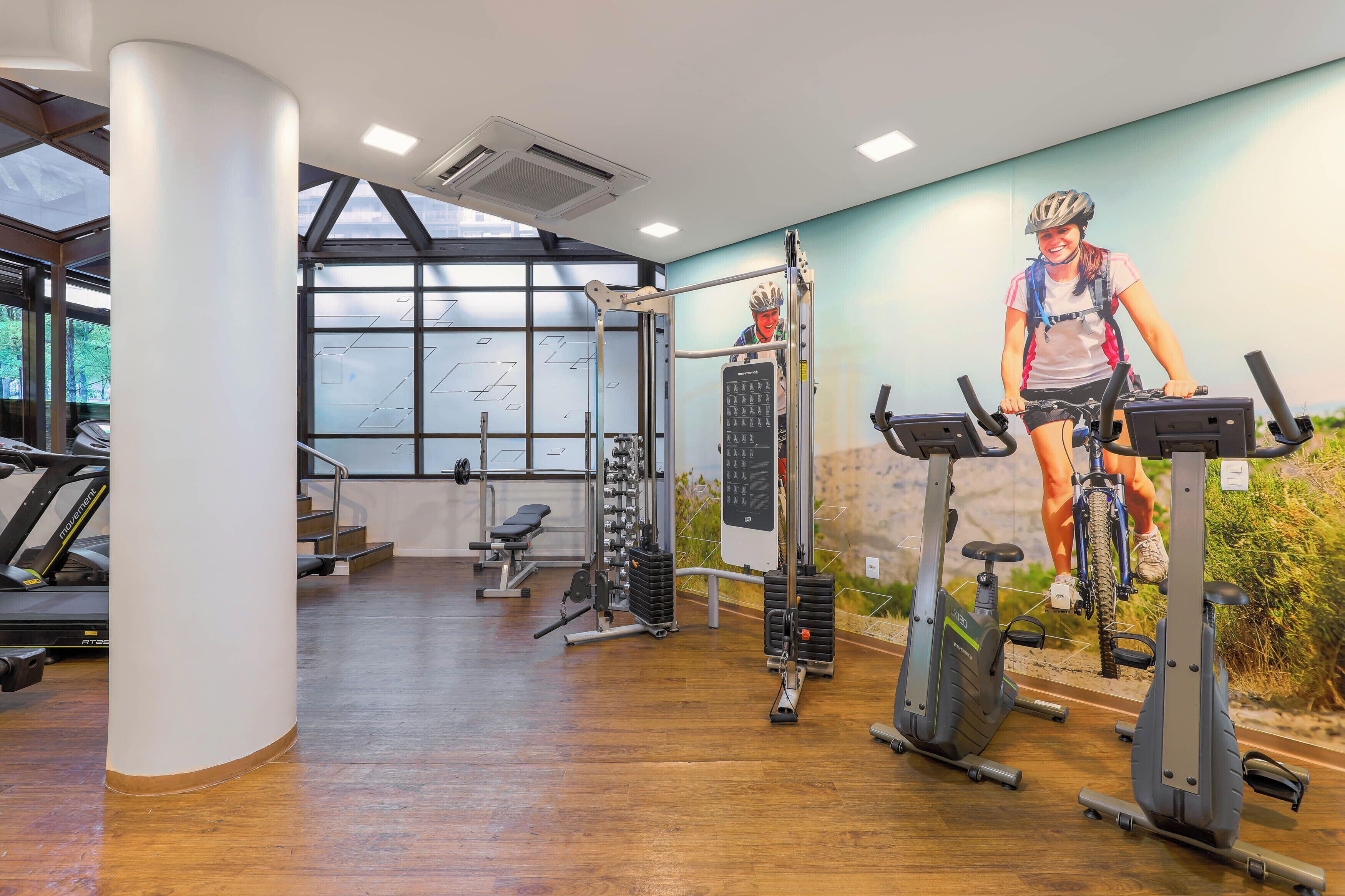 Un gimnasio con equipamiento de ejercicio, un mural y ventanas amplias.