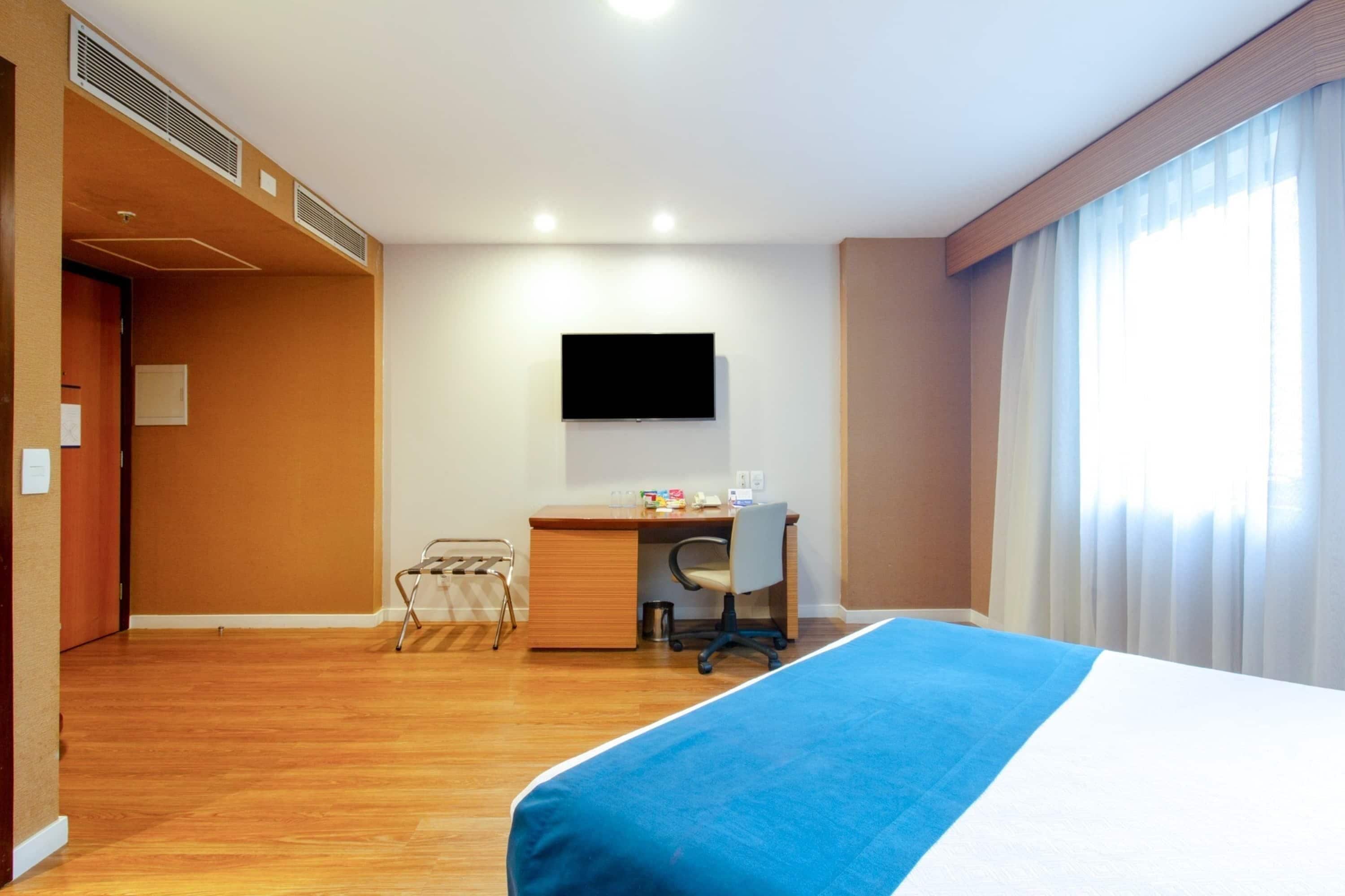 Quarto de hotel com cama, escrivaninha, cadeira e televisão fixada na parede.