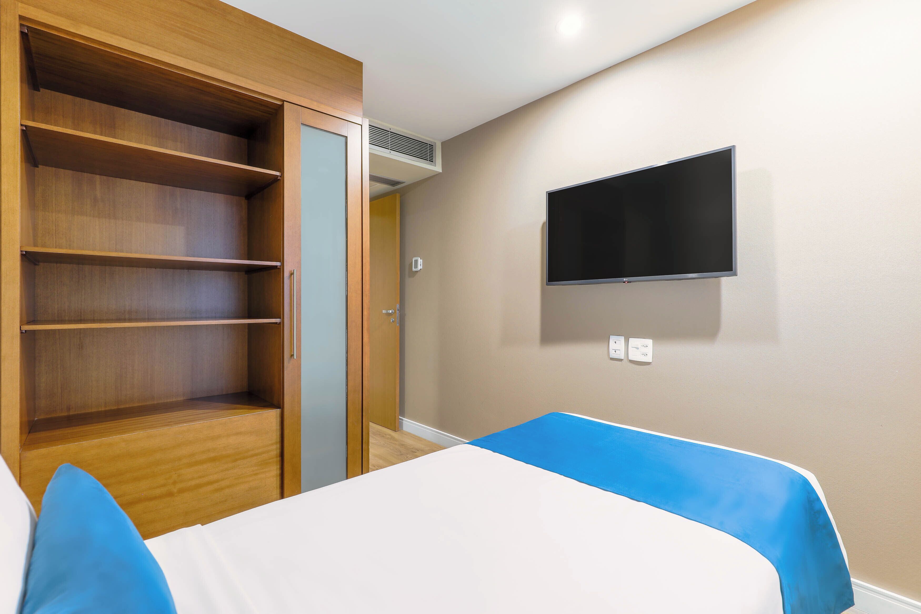 A hotel room with a wooden wardrobe, a flat-screen TV mounted on the wall, and two beds with blue bedspreads.