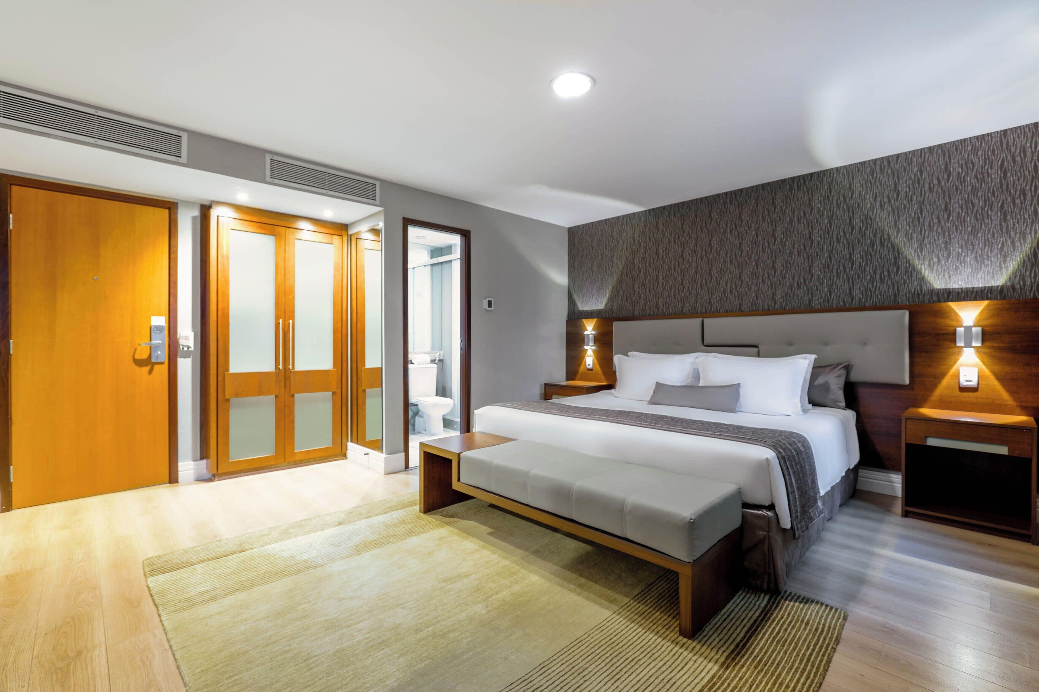 Una habitación de hotel moderna con una cama grande, un armario de madera y un baño visible a través de una puerta abierta.
