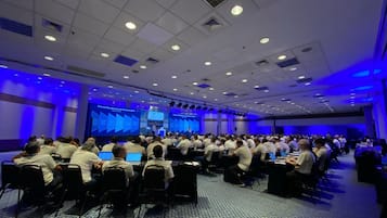 Sala de conferências com participantes sentados em mesas, olhando para uma tela de apresentação.