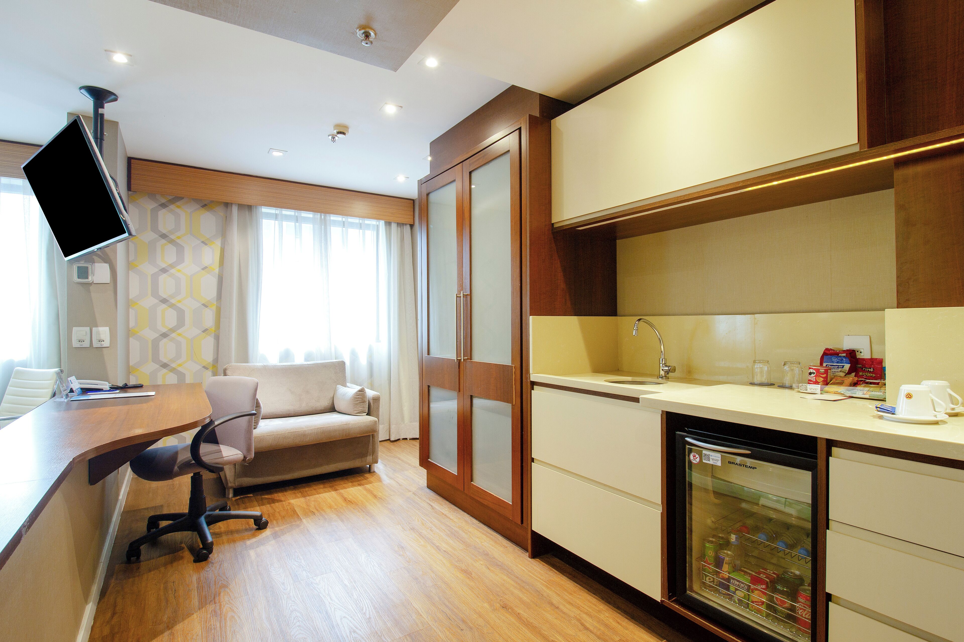 A compact hotel room with a kitchenette, a sofa, a desk with a computer, and a television.