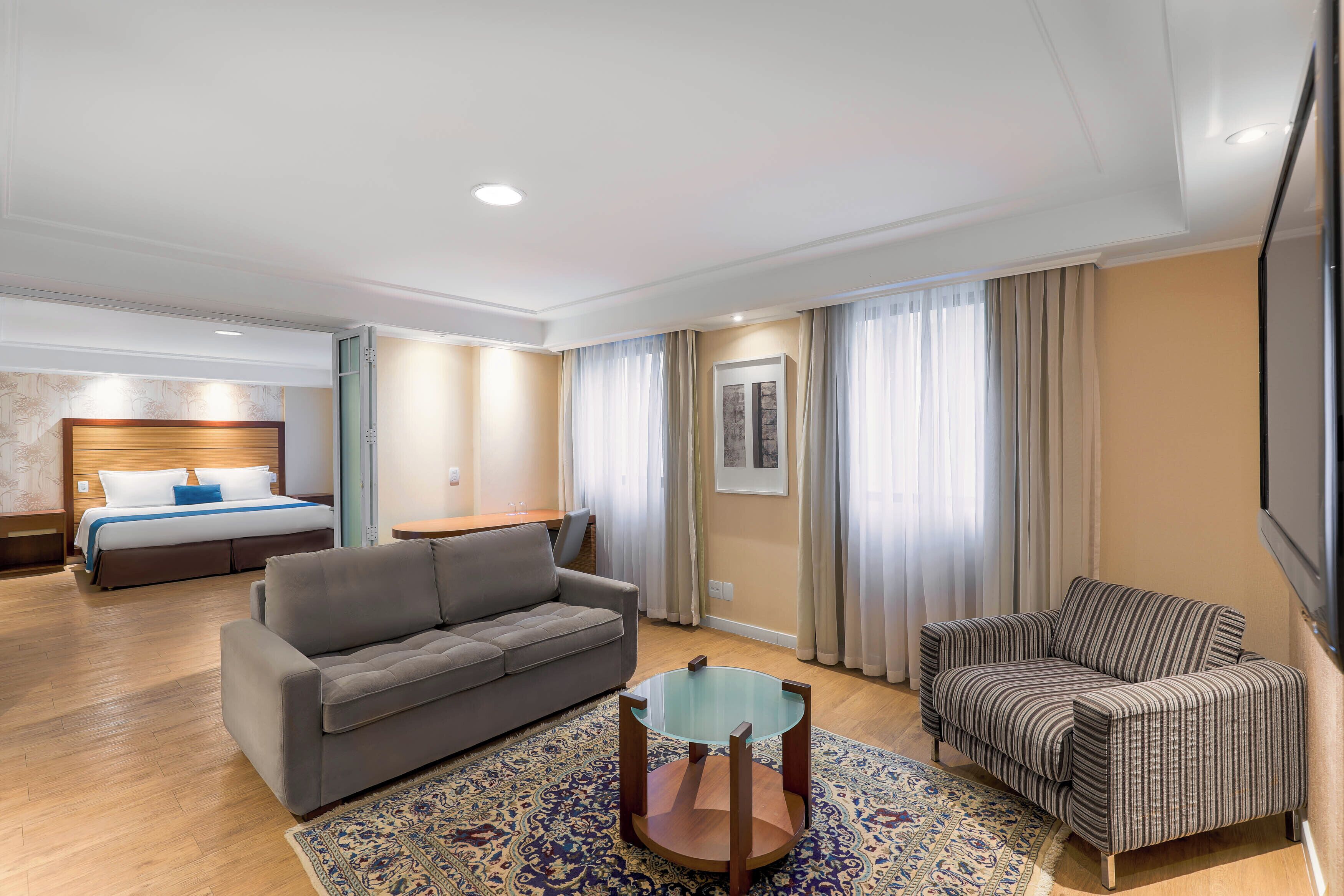 Habitación de hotel con cama, dos sofás, una mesita de vidrio, alfombra estampada y un ventanal con cortinas.