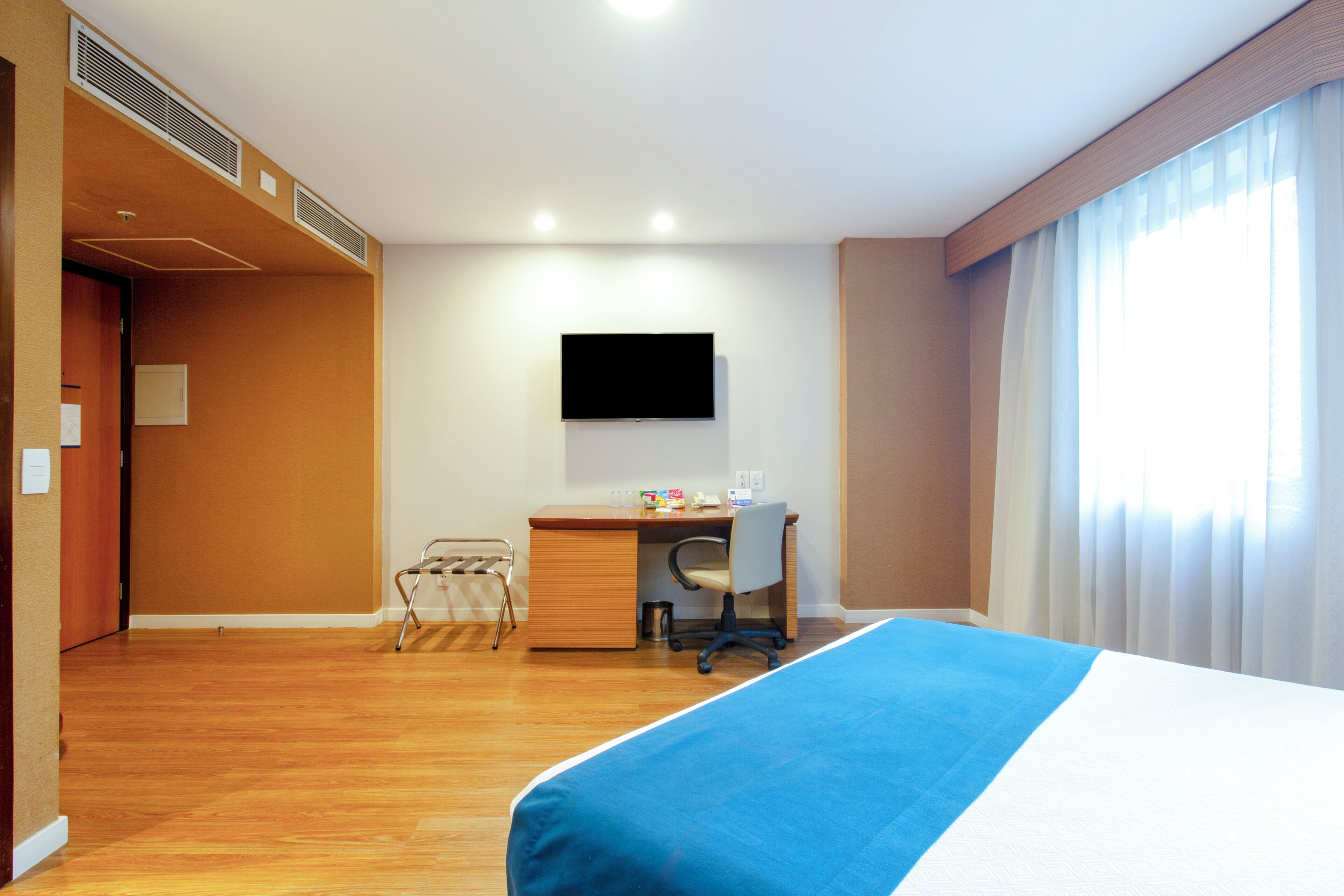 Una habitación de hotel con cama, escritorio, silla, televisor y ventana con cortinas.