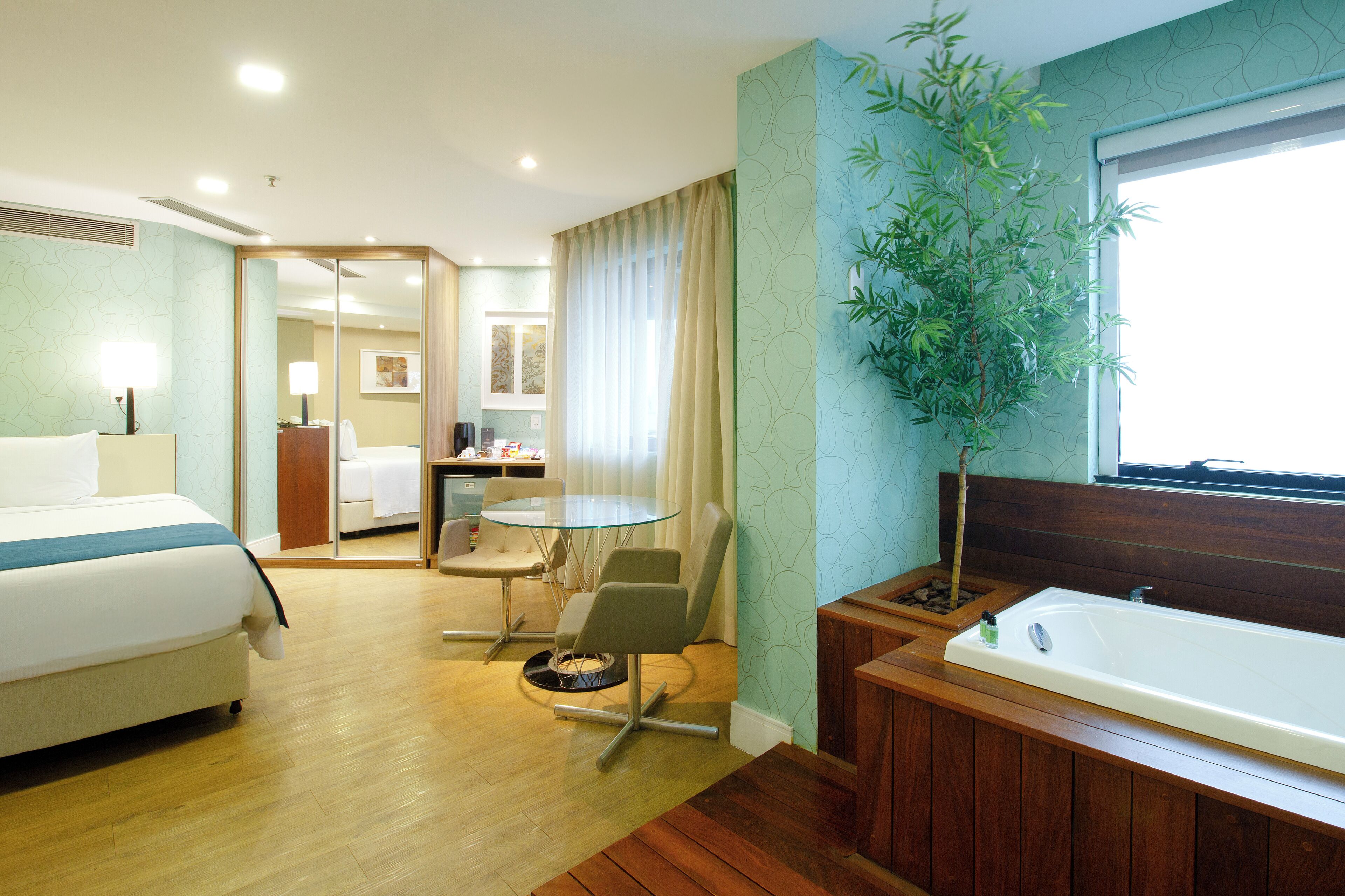 Una habitación de hotel con cama, una mesita redonda pequeña, una silla, bañera y una planta en maceta.