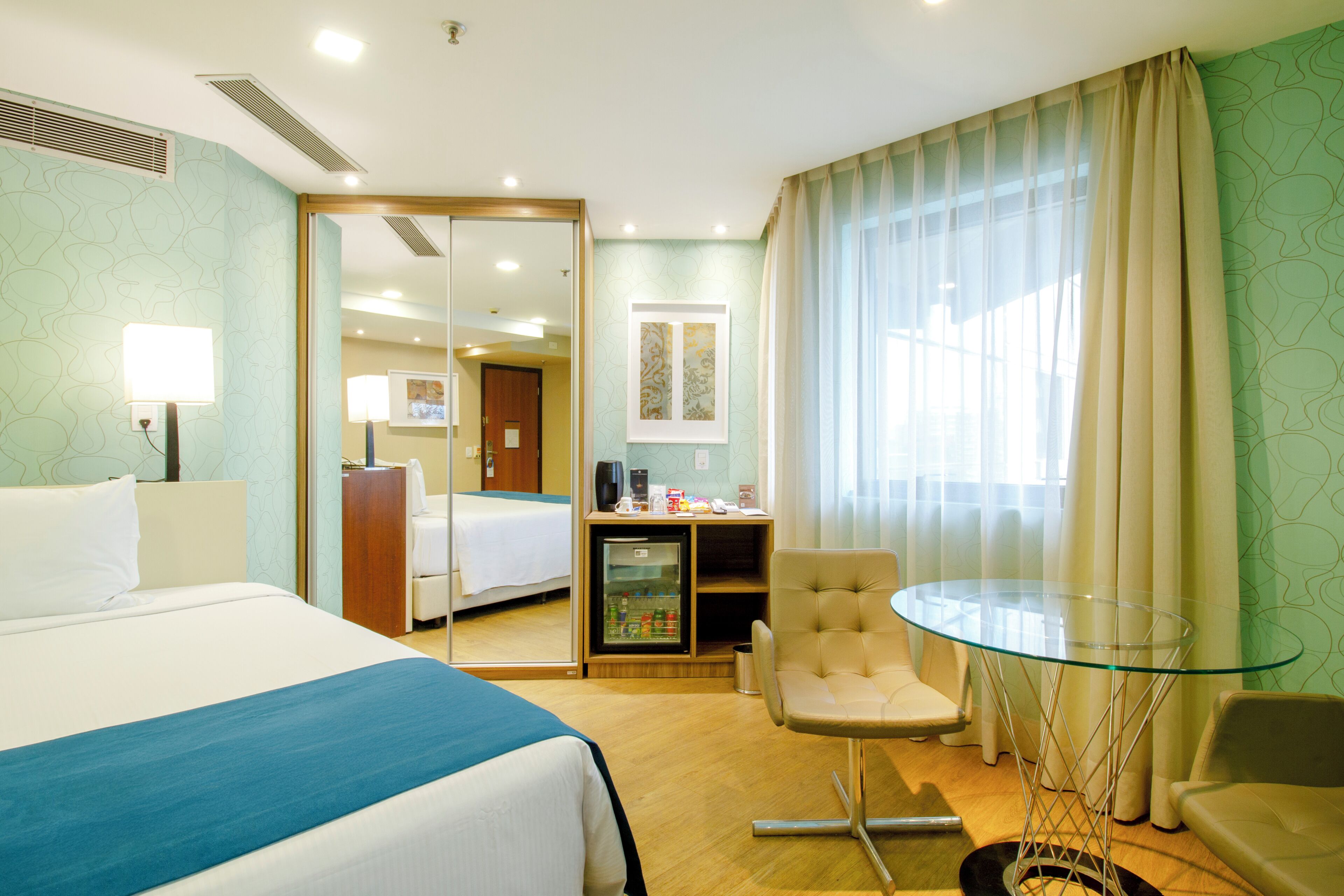 A hotel room with a bed, a desk, a chair, a glass table, and a minibar.