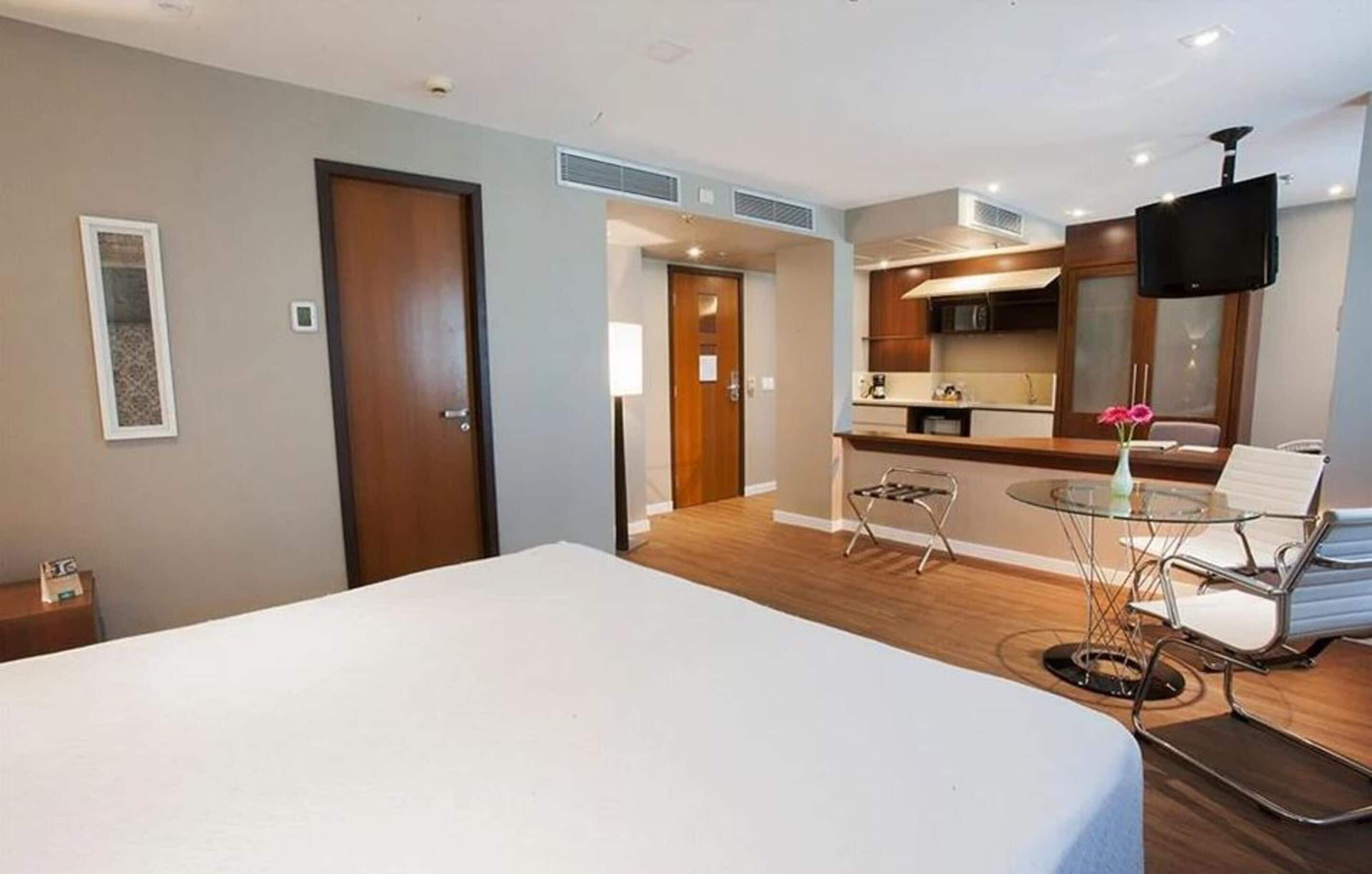 A hotel room with a bed, a small table with chairs, a kitchenette, and a TV.