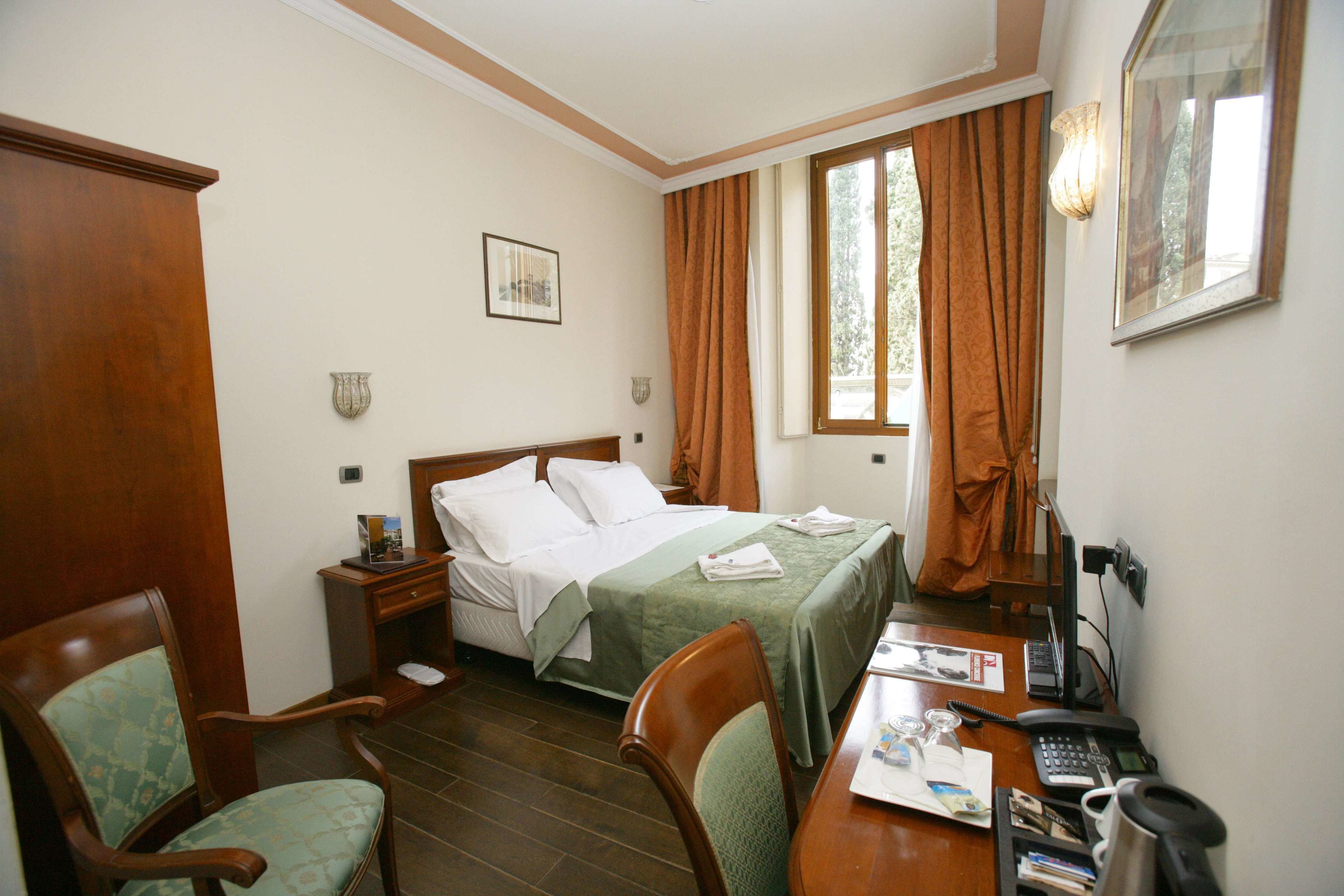 A hotel room with a bed, two chairs, a desk with a telephone and papers, and a window with curtains.