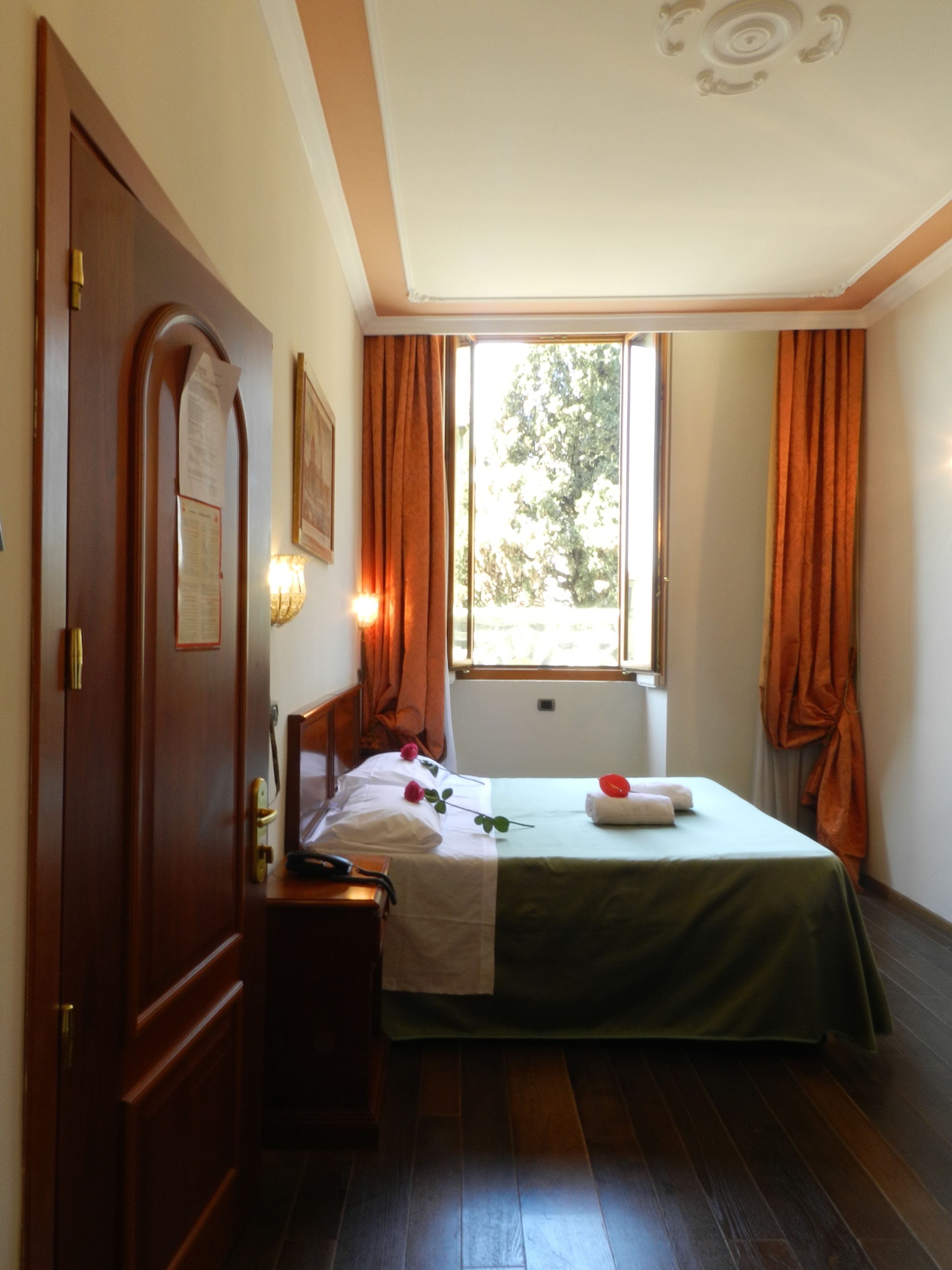Una habitación con cama, escritorio, ventana con cortinas, puerta y lámpara fijada a la pared.