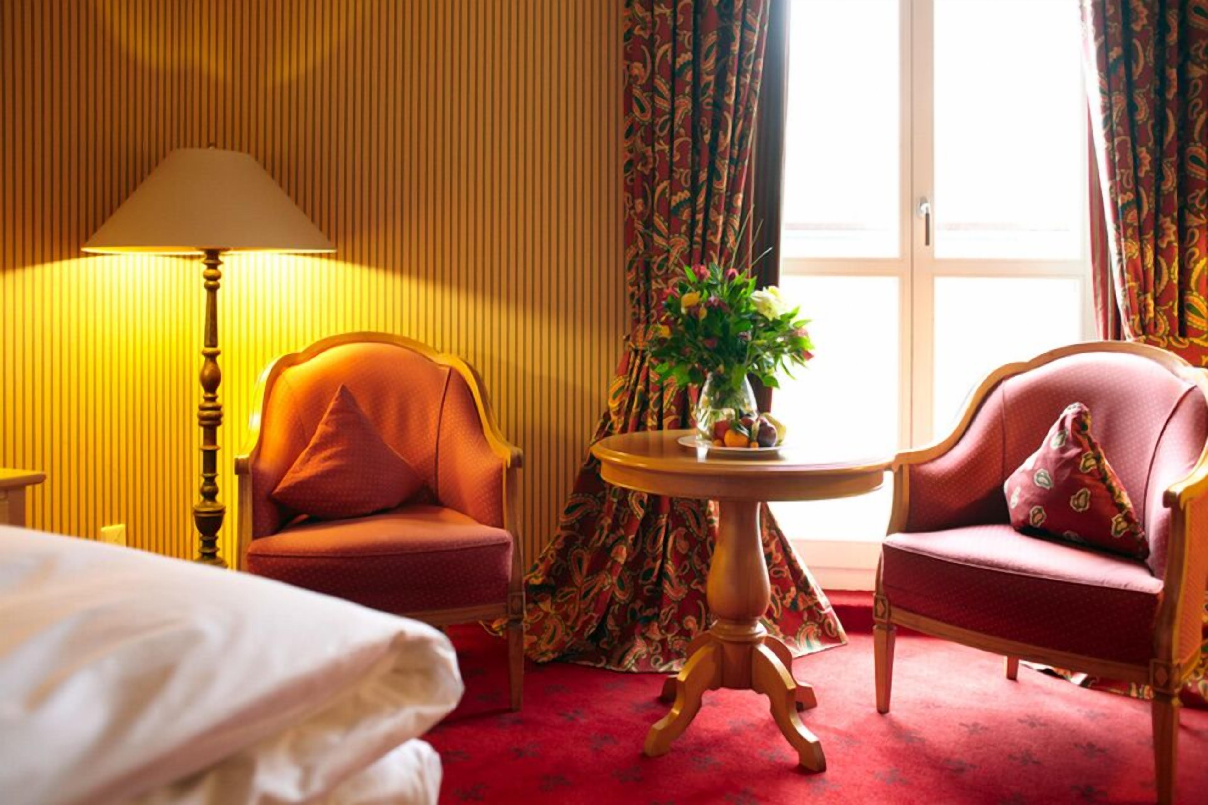 Habitación de hotel con cama, dos sillones, una mesa redonda con una planta, lámpara y ventana con cortinas.