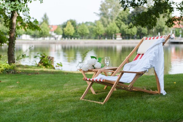 A wooden lounge chair with a white towel and striped cushion, positioned on a grassy area near a body of water.