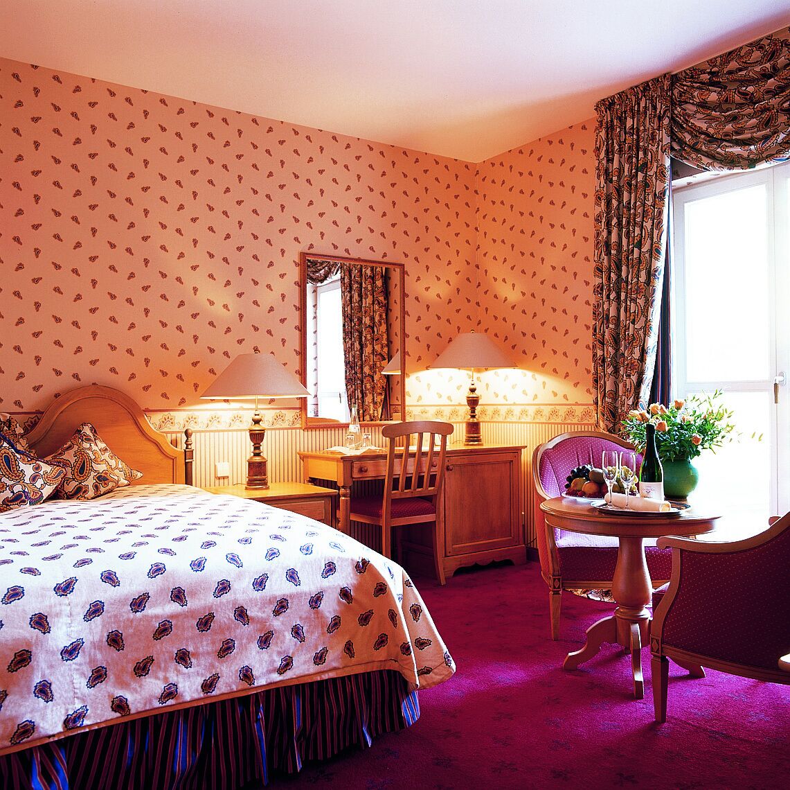 Ein Hotelzimmer mit einem Bett, einem Schreibtisch mit Stuhl, einem Tisch mit Stuhl, einem Spiegel und einem Fenster mit Vorhängen.
