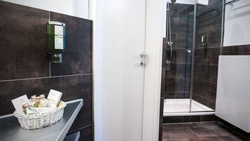 Un baño moderno con ducha, lavamanos y un cesto de artículos de aseo, además de un dispensador de jabón en la pared.