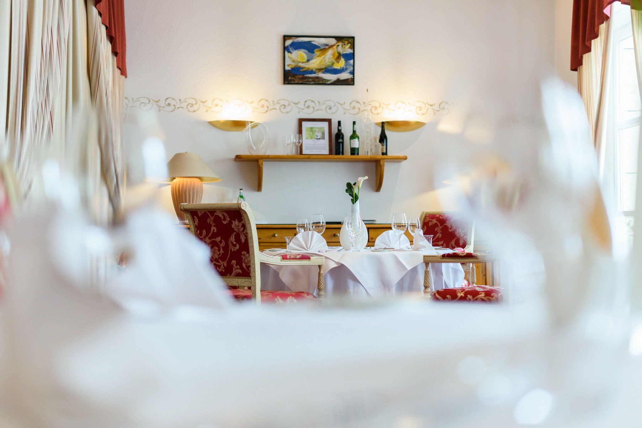 A dining area with tables set for a meal, a wine rack above the tables, and a painting on the wall.