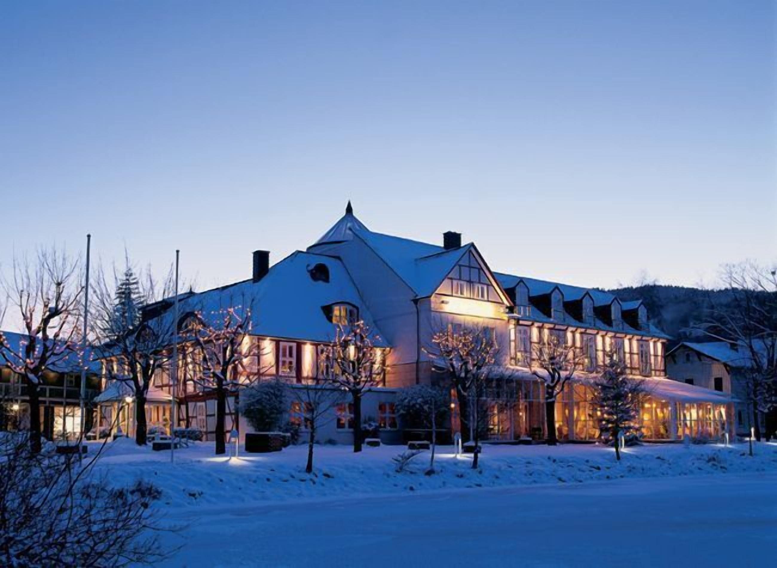 A large, multi-story building with a steeply pitched roof, likely a hotel or resort, surrounded by snow-covered ground and bare trees.