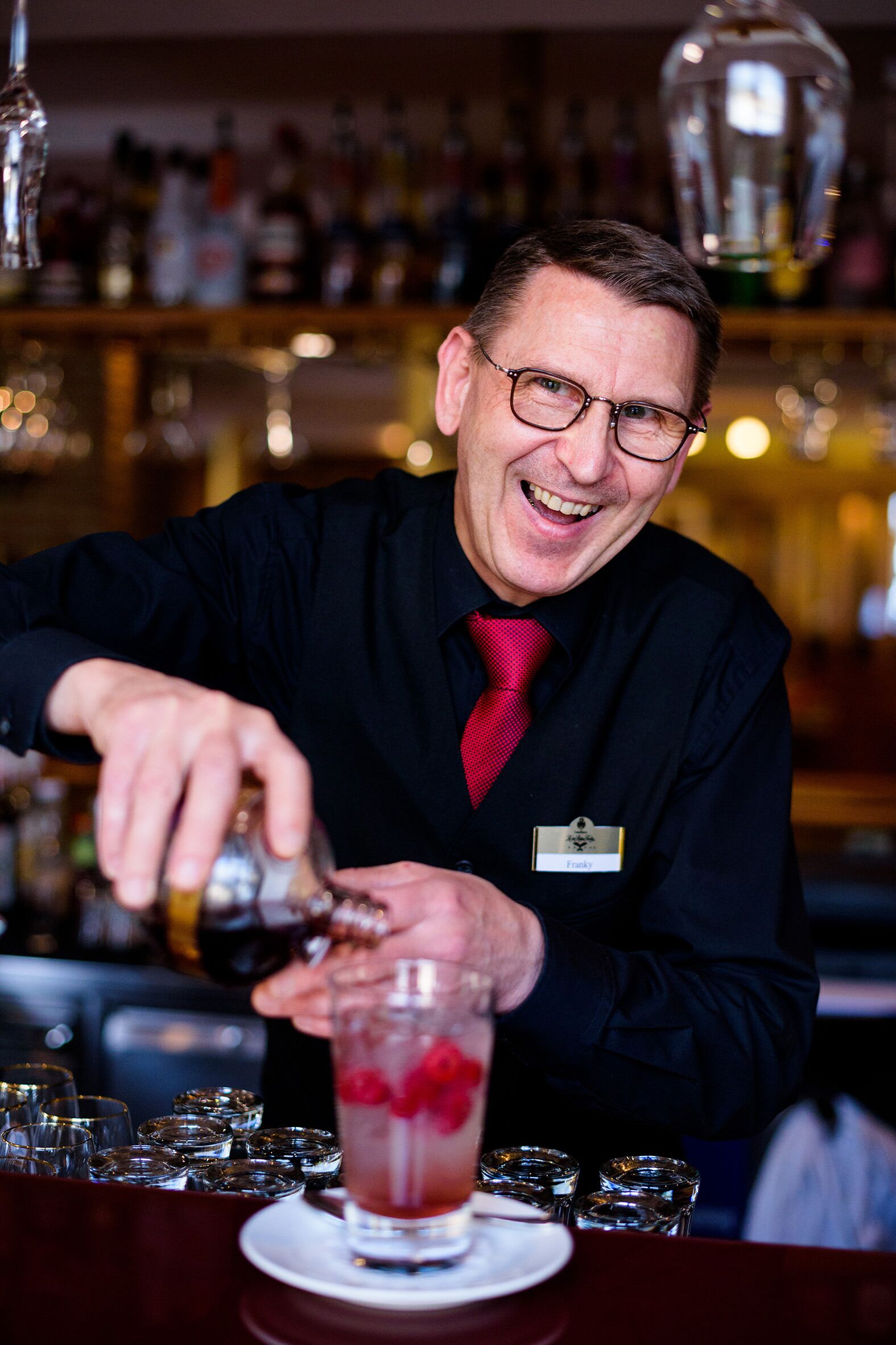 Un bartender preparando una bebida en la barra, con una estantería repleta de botellas al fondo.