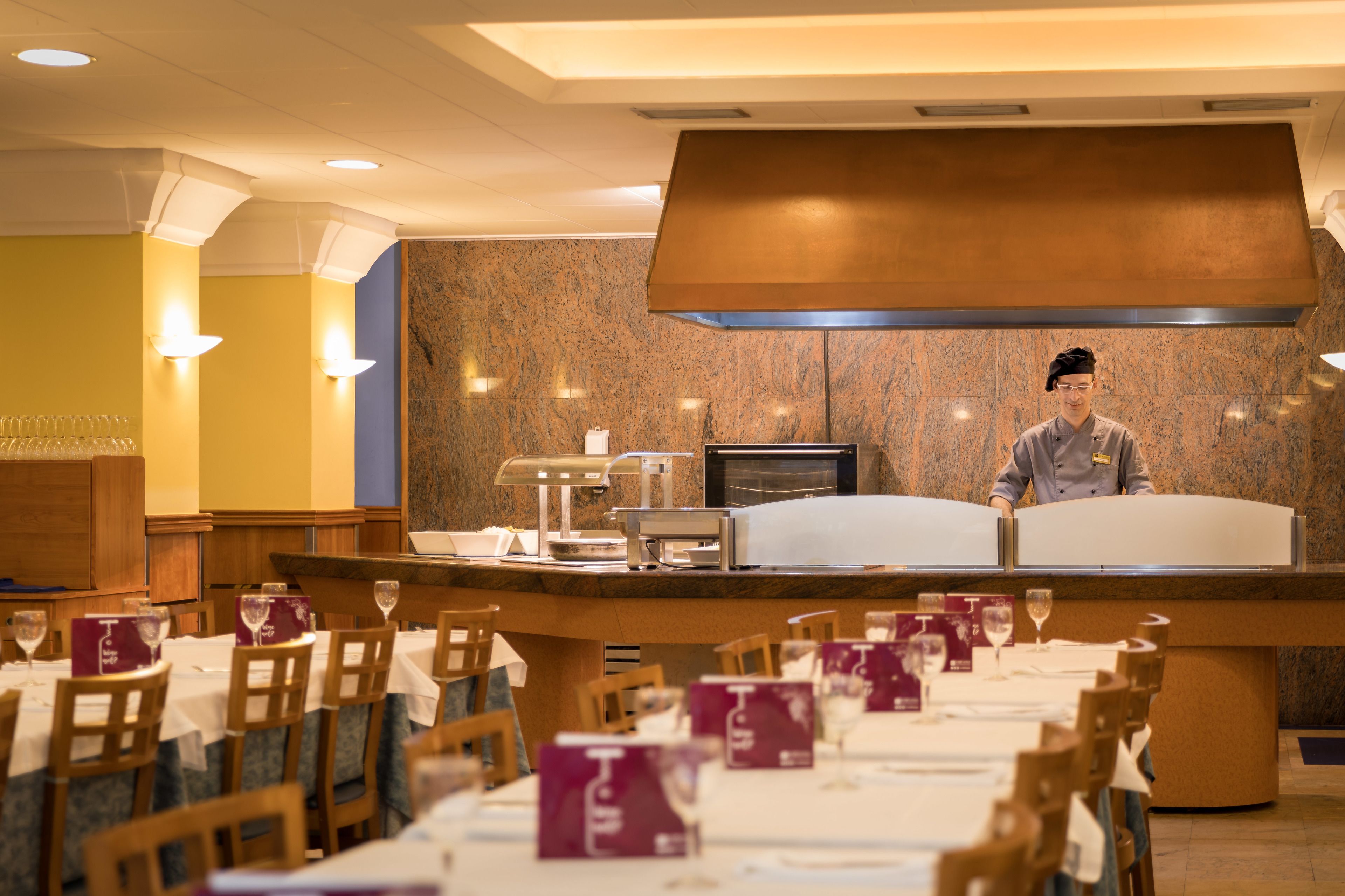 Un restaurante con un chef detrás de la barra, mesas con menús y una pared de mármol al fondo.