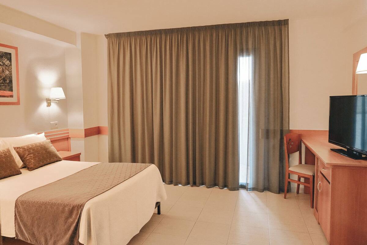 Una habitación de hotel con una cama, un escritorio con televisor y un ventanal con cortinas.