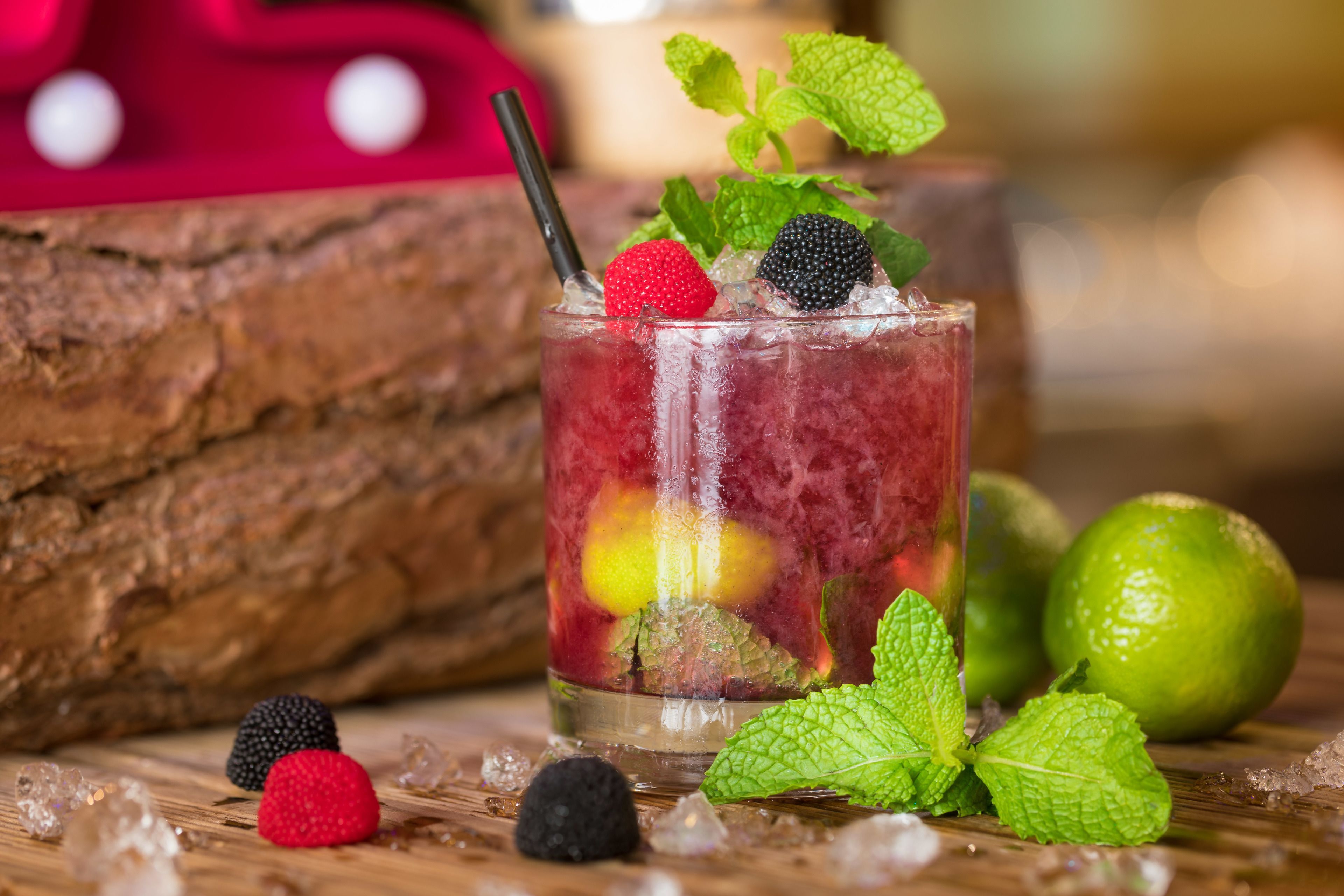A glass of iced beverage with berries and mint, surrounded by ice cubes and lime on a wooden surface.