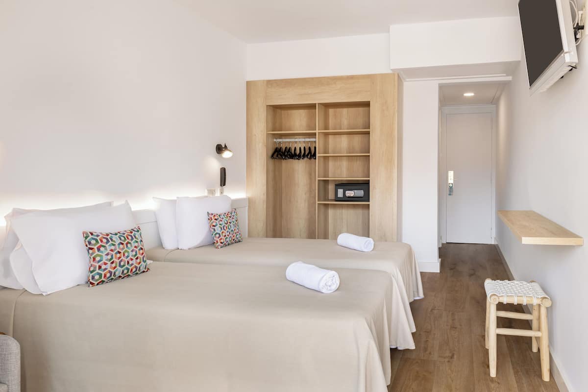 Habitación de hotel con dos camas, un armario de madera, un taburete pequeño de madera y un televisor fijado a la pared.