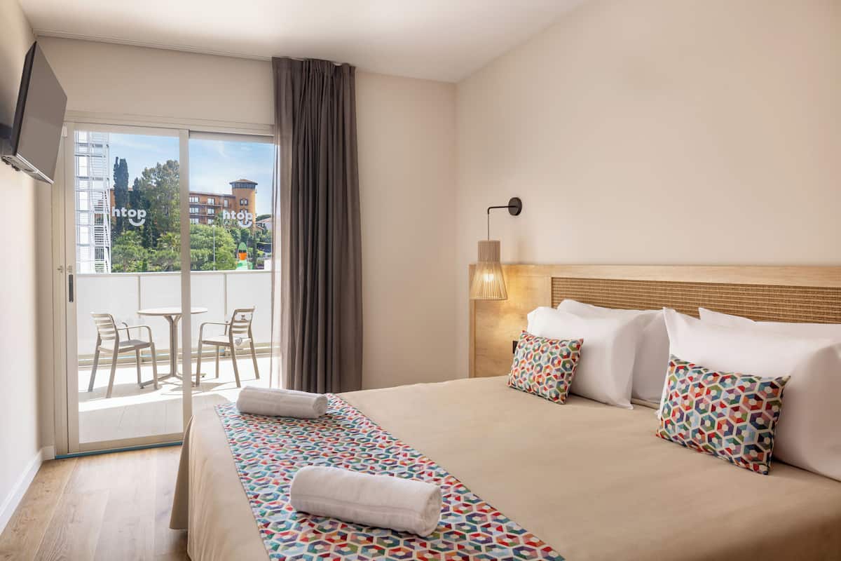 Habitación de hotel con cama, balcón con sillas y vista a la ciudad.