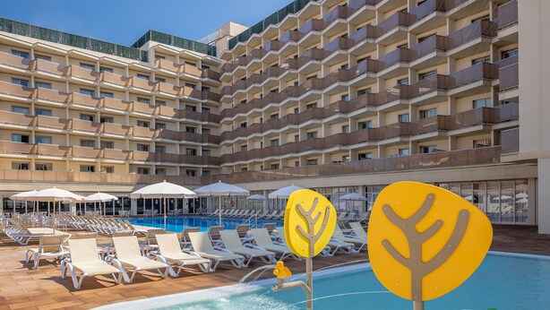 Zona de piscina hotelera con tumbonas y sombrillas, rodeada por un edificio de varios pisos con balcones.