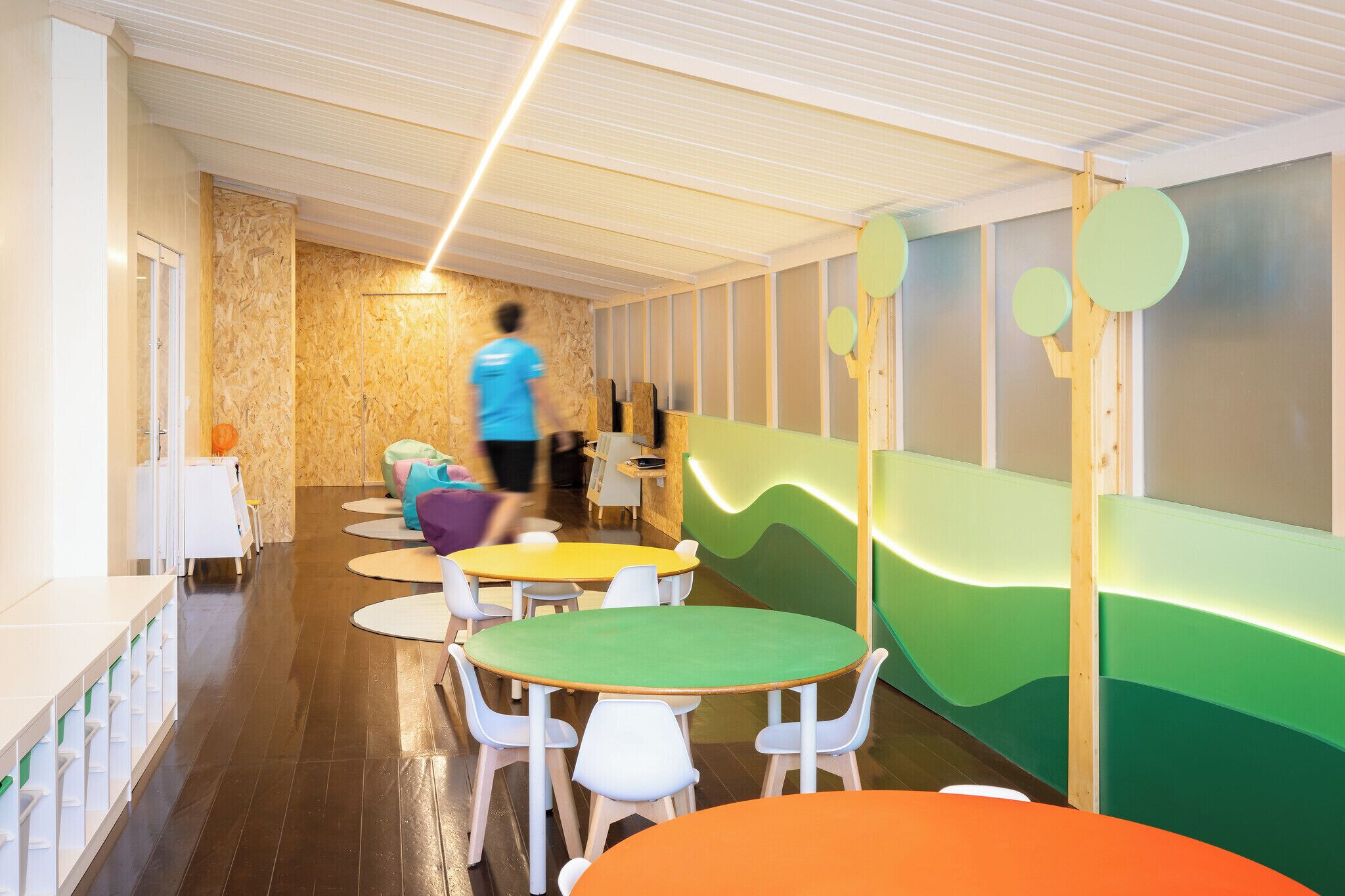 Zona de juegos infantiles con mesas y sillas de colores, una estructura de madera y una pared con diseños abstractos.