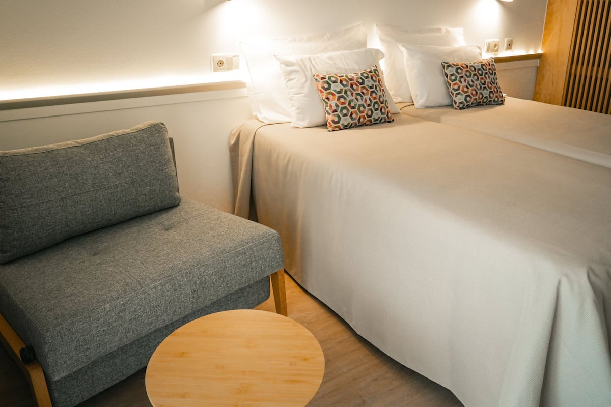 Un dormitorio con un sofá gris, una mesa redonda de madera y dos camas con almohadas estampadas.