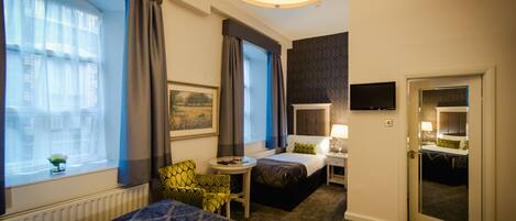 A hotel room with a bed, a desk, a chair, a small table, a window with curtains, a painting, a television, and a bathroom mirror.