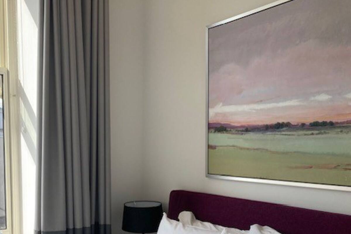 A hotel room with a large bed, a headboard, a painting of a landscape, a window with curtains, and a bedside table with a lamp.