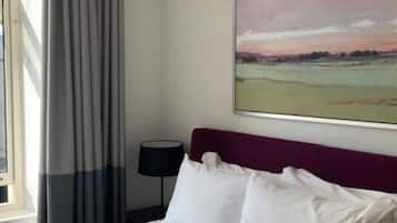 Habitación de hotel con una cama grande, cabecera, cuadro de un paisaje, ventana con cortinas y mesita de noche con lámpara.