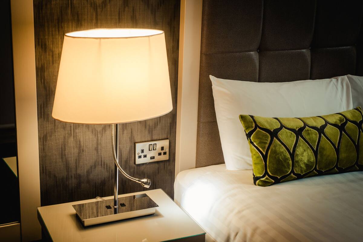 A neatly made bed with white linens and a green patterned pillow, a bedside lamp, and a power outlet on the nightstand.