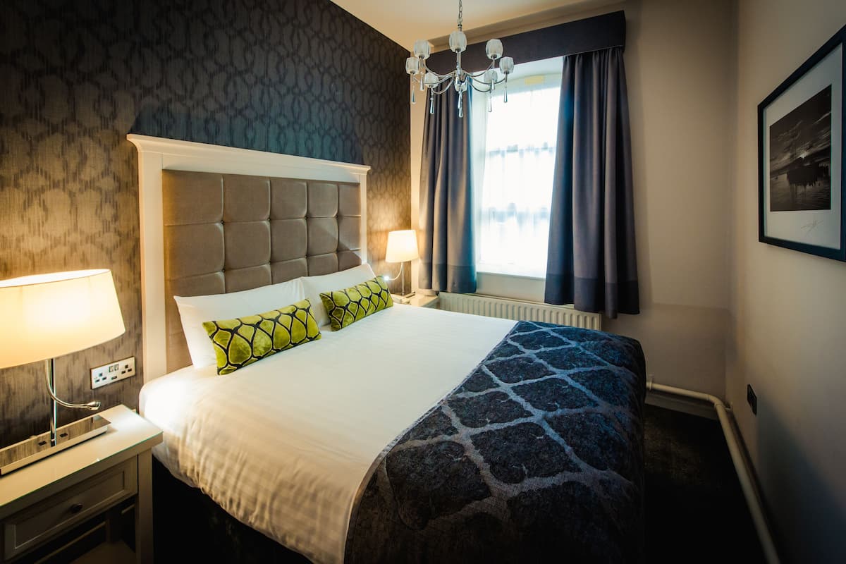 A hotel room with a large bed, a nightstand with a lamp, a chandelier, and a window with curtains.
