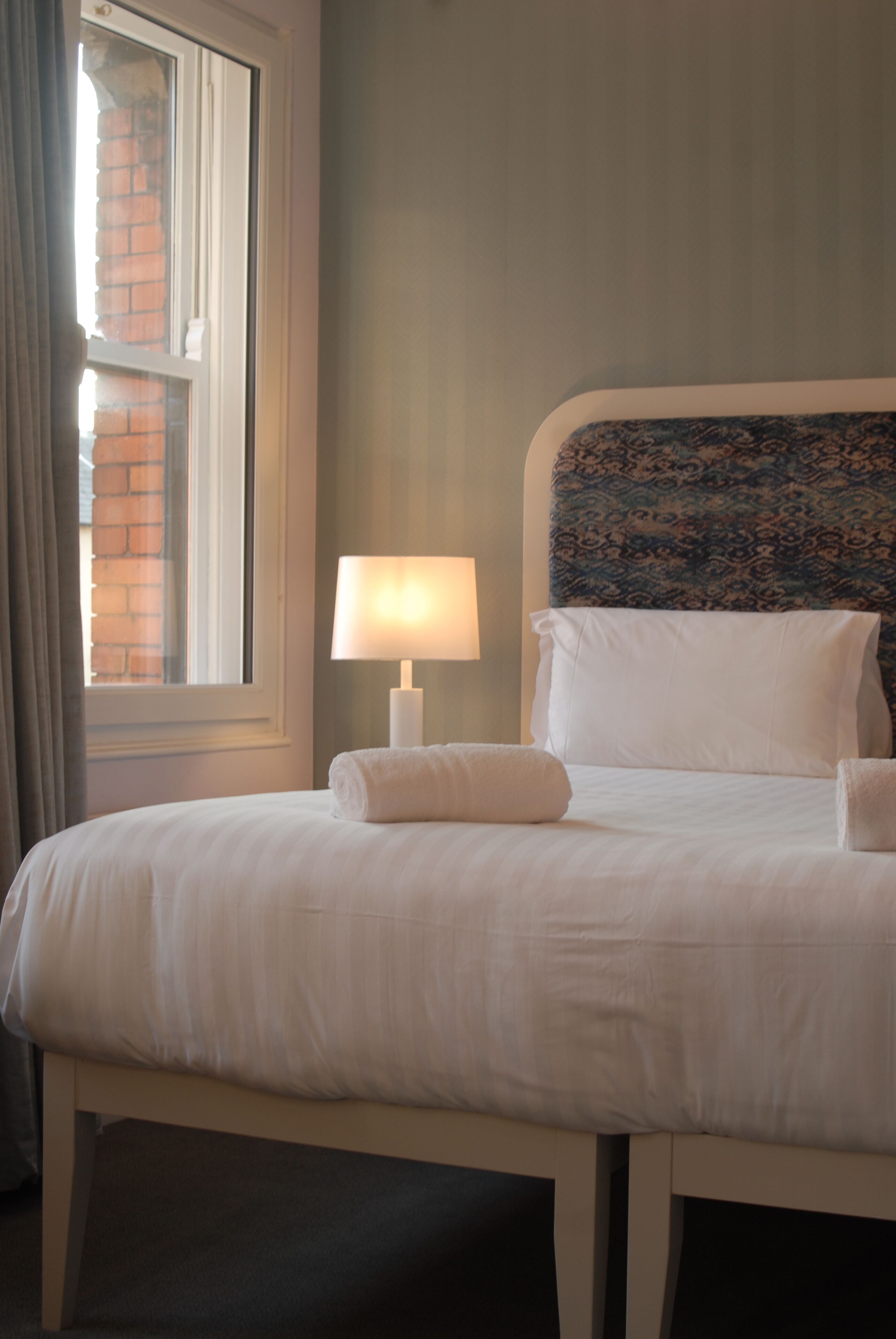 A neatly made bed with white linens, a bedside lamp, and a window with curtains.