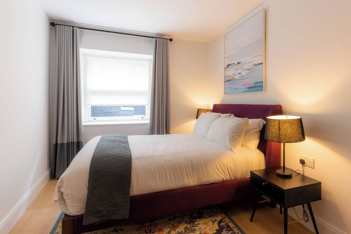 A neatly made bed with a headboard, a bedside lamp, and a window with curtains.