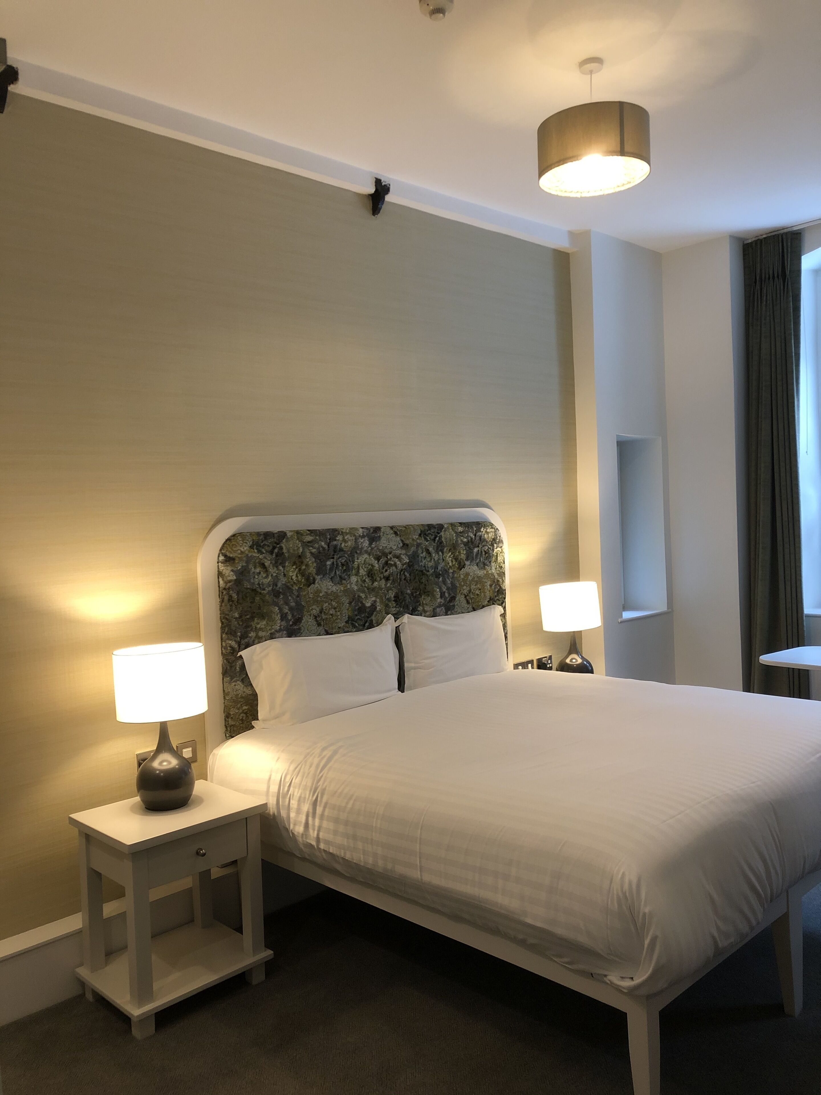 A neatly made bed with white linens, two bedside tables with lamps, a headboard with a floral pattern, and a window with curtains.