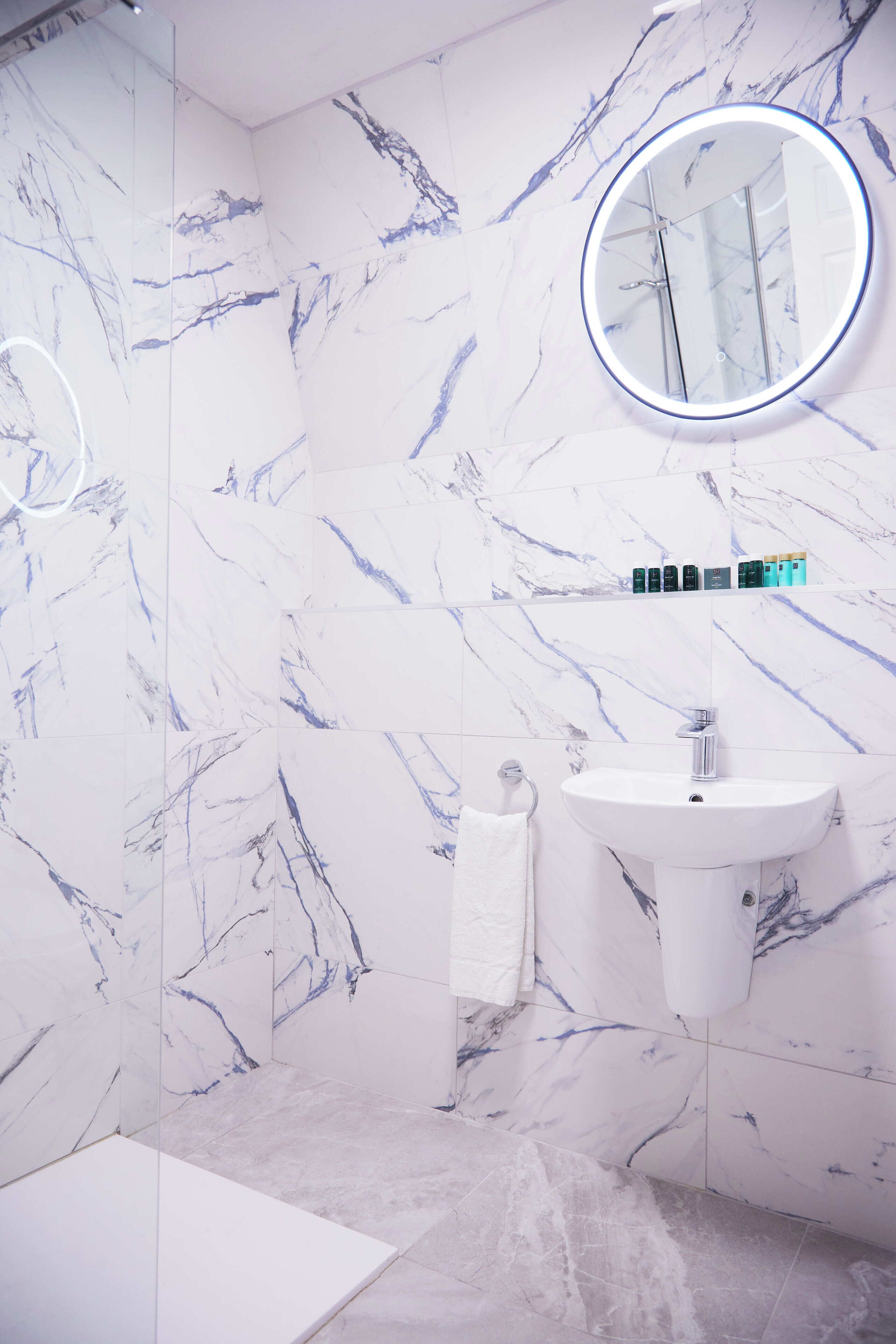 Un baño con pared de mármol blanco, un espejo redondo con luz, un lavabo blanco y un estante con artículos de aseo.