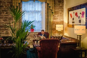 A cozy room with a stone wall, a leather sofa, a red armchair, a potted plant, and a window with curtains.