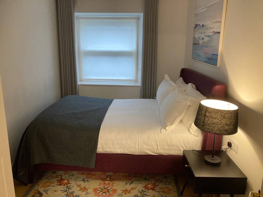 A neatly made bed with a grey blanket and white linens, a bedside table with a lamp, and a floral-patterned rug on the floor.