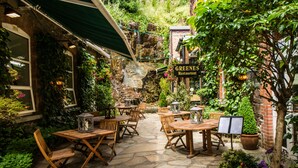 Uma área de pátio ao ar livre com piso de pedra, mesas e cadeiras de madeira, plantas em vasos e uma placa para o Restaurante Greenes.
