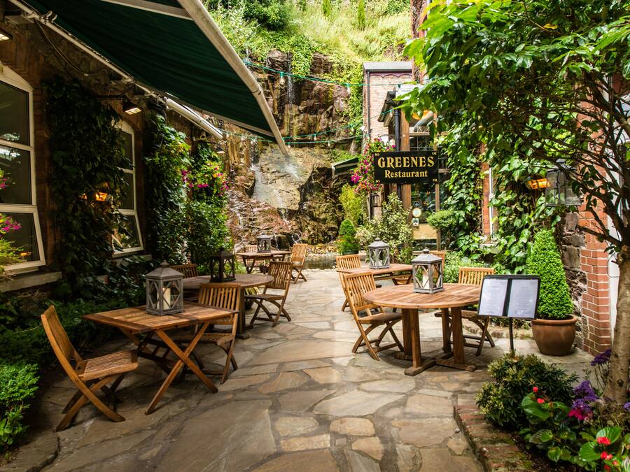Un patio al aire libre con suelo de piedra, mesas y sillas de madera, plantas en macetas y un letrero para el Restaurante Greenes.