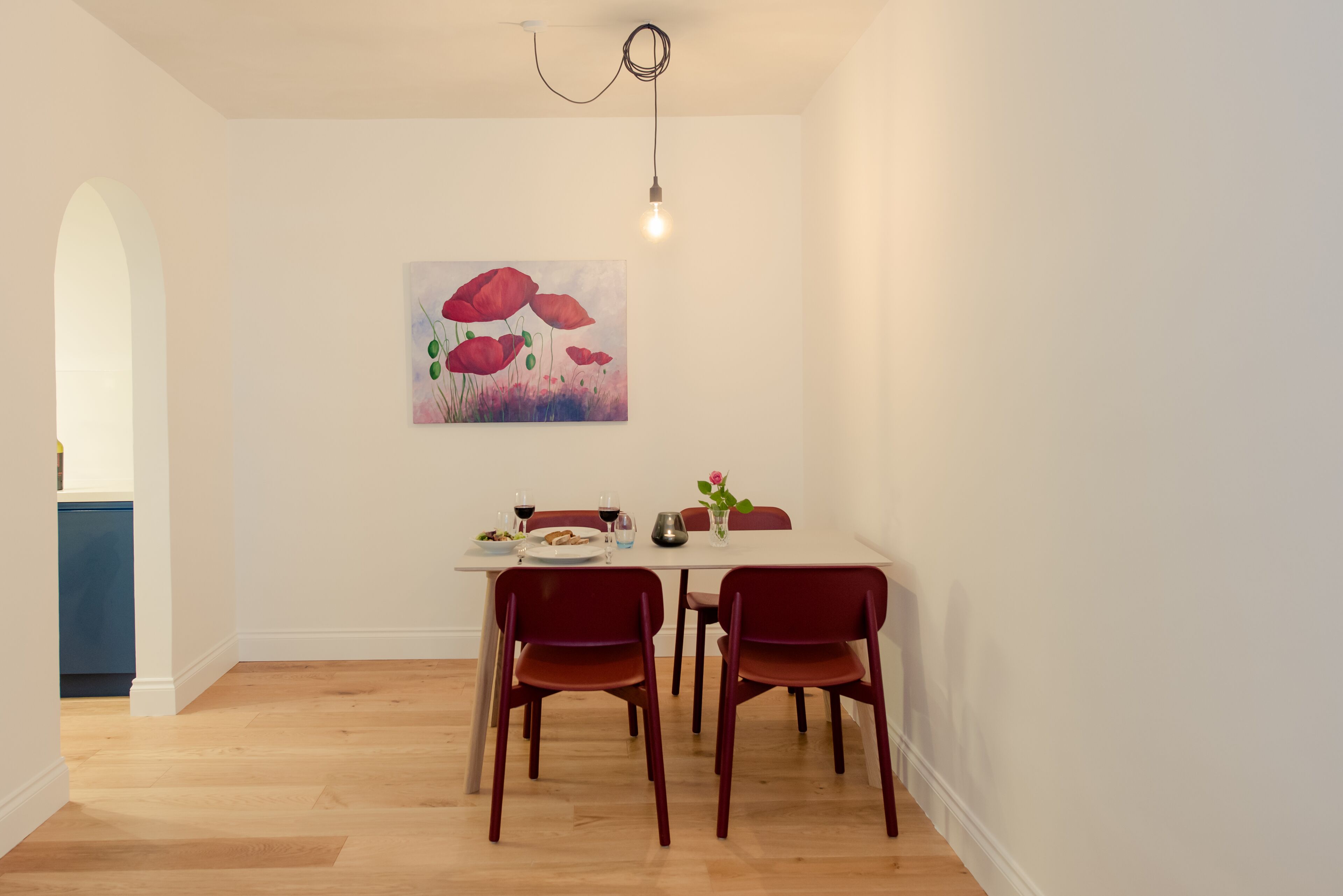 A dining area with a white table, four red chairs, a painting of red flowers on the wall, and a light fixture above.