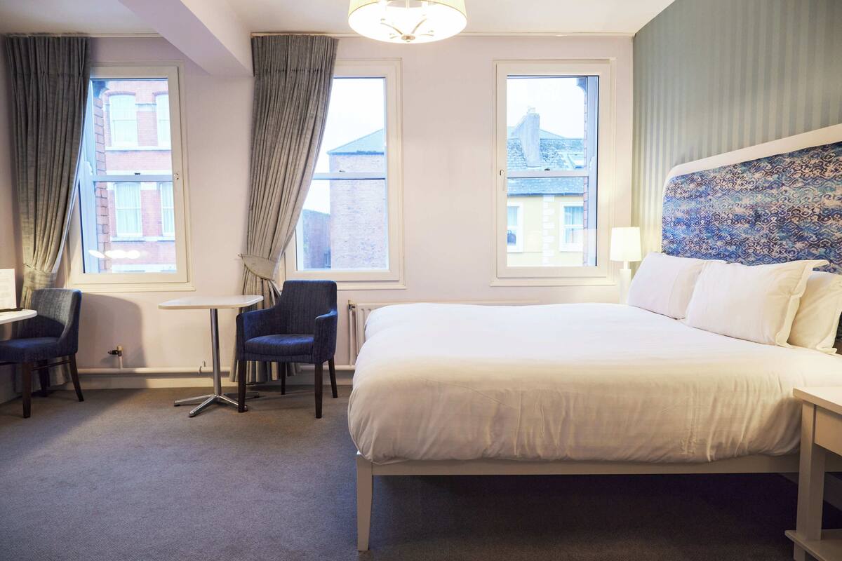 A hotel room with a large bed, two chairs, a small table, and two windows with curtains.