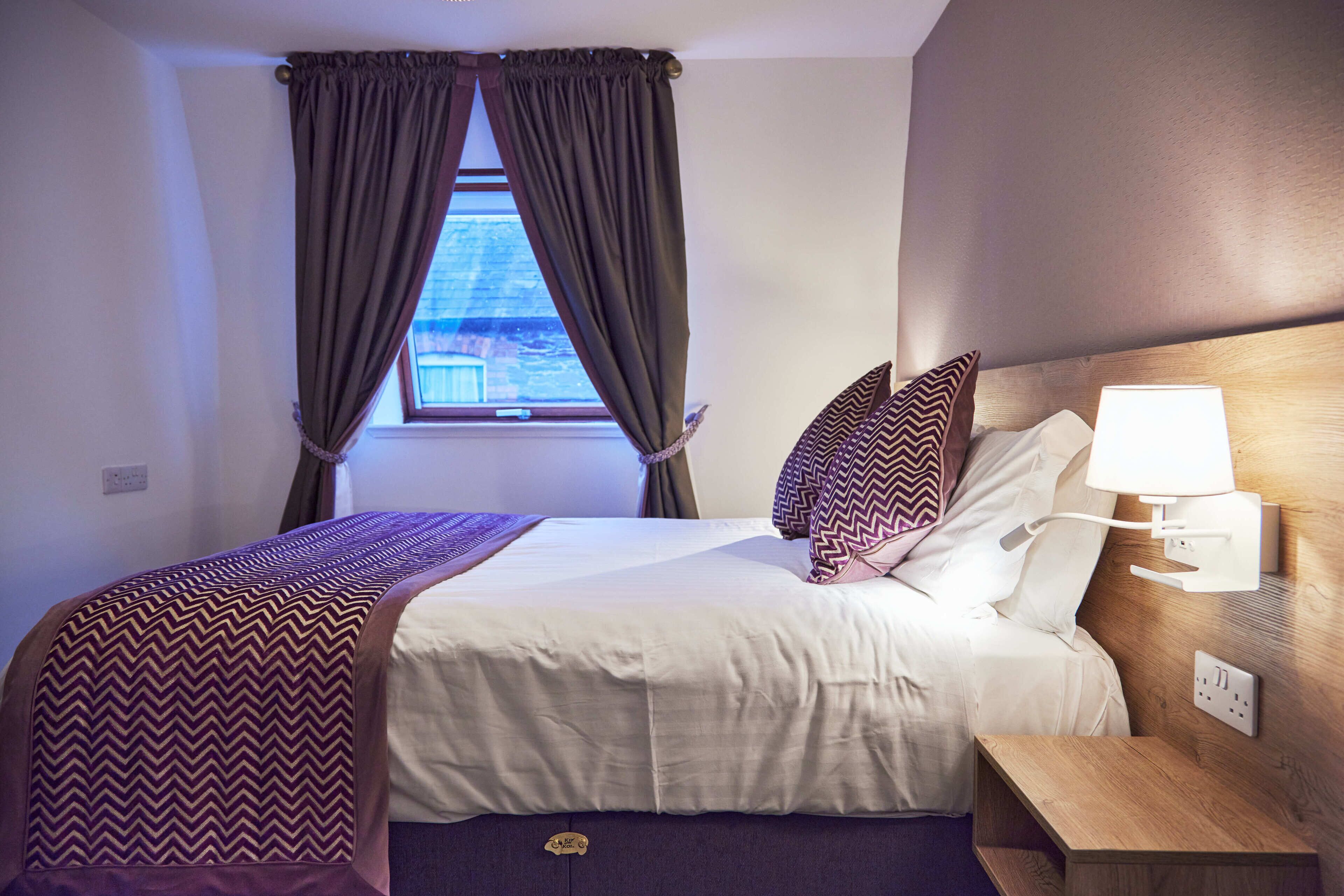 Habitación de hotel con cama, mesita de noche, lámpara y ventana con cortinas.