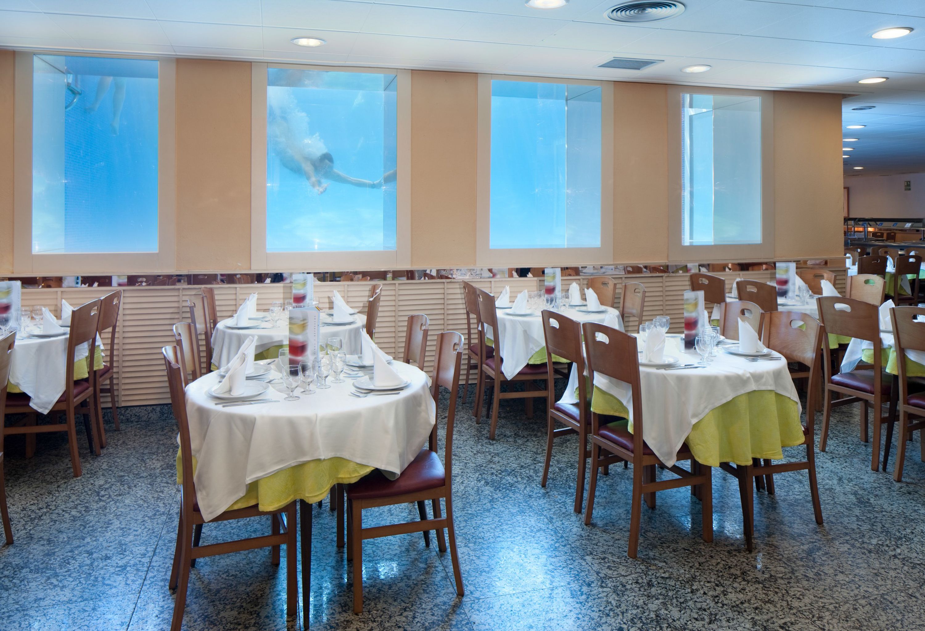 A dining area with multiple tables and chairs, large windows with a view of a swimming pool, and a tiled floor.