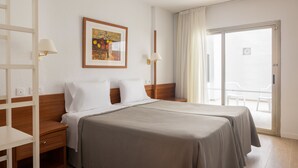 Habitación de hotel con cama doble, mesita de noche, lámpara, un cuadro en la pared y puerta corrediza de vidrio al balcón.